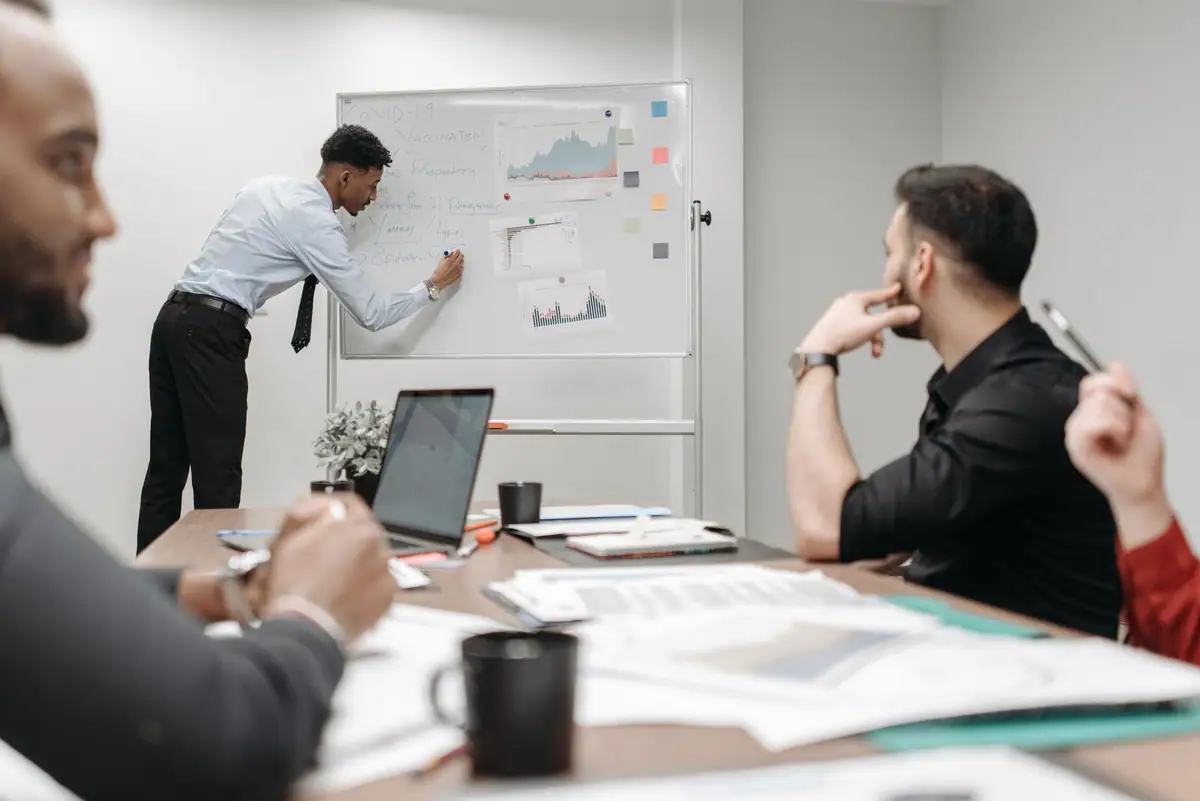 Presenter sketching charts on a whiteboard while colleagues follow along at a meeting table